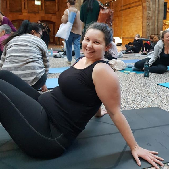 A photo of Sarah in a relaxed seated position on her yoga mat in the Hintze Hall in the Natural History Museum. Sarah is smiling at the camera.